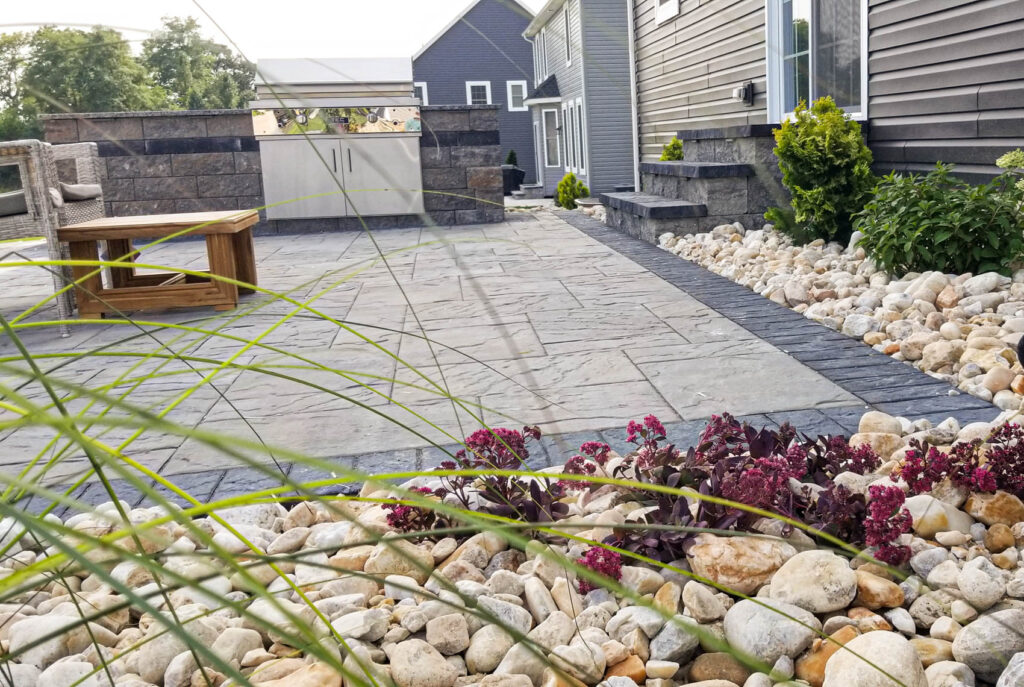 Patio featuring stone pavers, outdoor kitchen area with grill, and landscaped border of rocks and plants, illustrating hardscaping design for enhancing outdoor spaces.